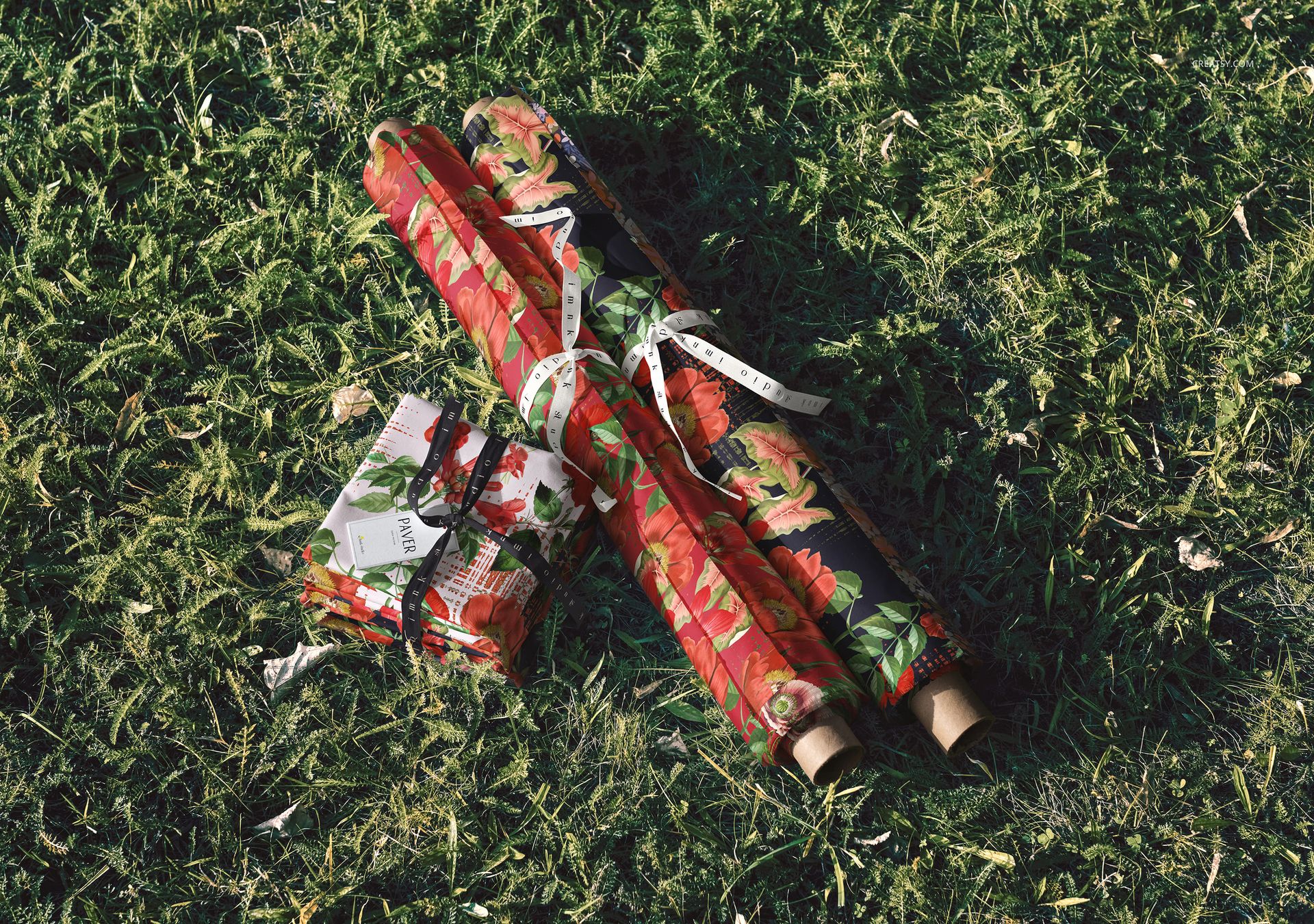 PSD mockup featuring patterned fabric rolls and stacks arranged outdoors on lush green grass, viewed from above.