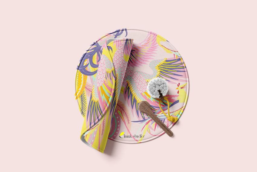 Round polyester bath mat mockup featuring a colorful bird and feather pattern in pink, yellow, and purple, shown with a wooden brush and a rolled towel on a light background.