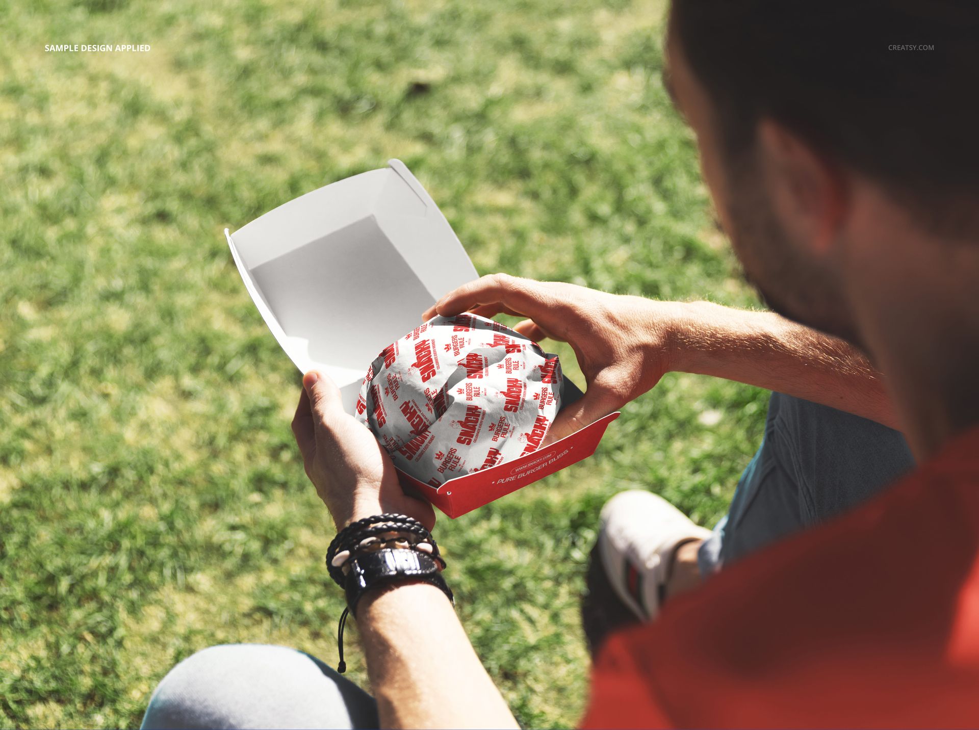 Person holding a burger wrapped in printed paper inside a red and white box, sitting outdoors on green grass.
