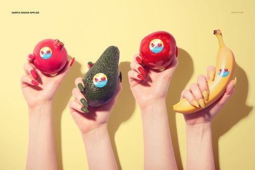 PSD mockup featuring pomegranate, avocado, and banana, each with a round sticker, held by hands on a yellow surface.