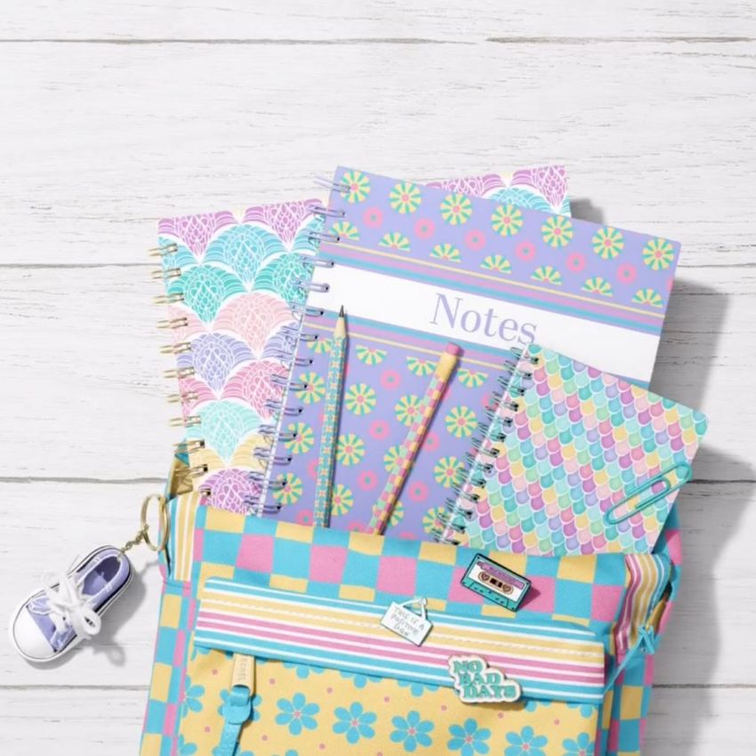 School backpack mockup with spiral notebooks in pastel colors and scallop patterns, shown on a light wooden surface.