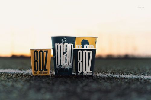 Four paper cups with bold graphic designs stand on a sports field, captured from a low front angle at sunset.