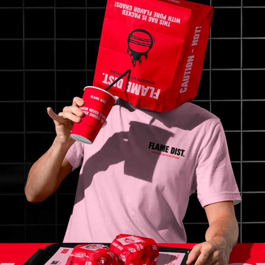 Man wearing a red paper bag with printed text on his head sits at a table with fast food packaging mockup in bold red colors.