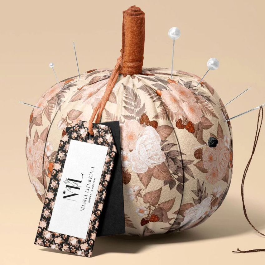 Fabric pumpkin mockup with a beige and brown floral pattern, used as a pincushion with pins and a tag attached, on a neutral background.