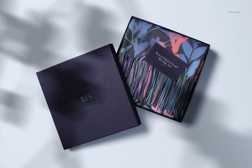 Woven fringed scarf with colorful botanical pattern displayed inside an open gift box, top view on light background.