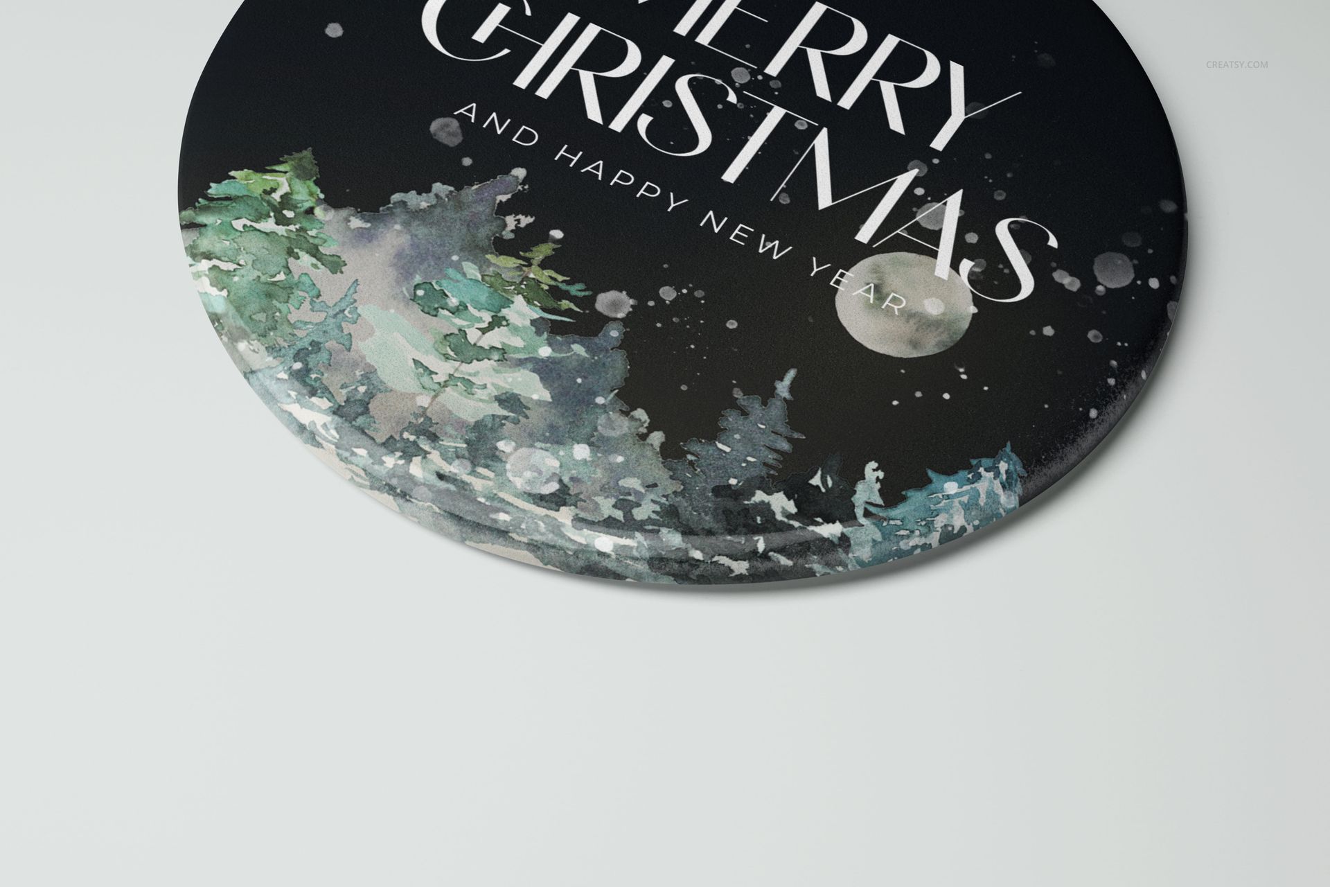 Round ceramic Christmas ornament mockup with dark background, white text, and watercolor trees, front angled view.