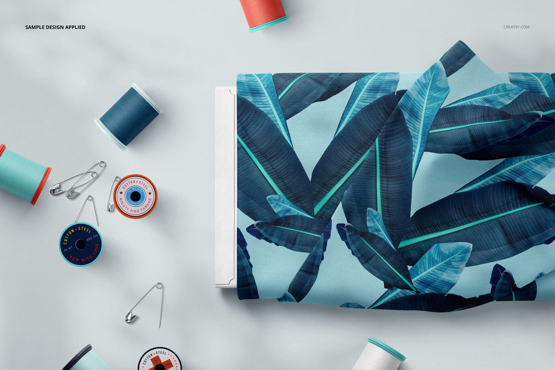 Fabric bolt featuring a tropical blue leaf design, placed next to assorted thread spools and safety pins in a mockup scene.