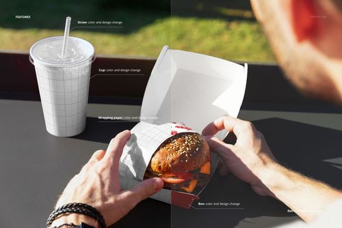PSD mockup showing hands holding a burger in a white packaging box, with a drink cup placed beside on a dark surface.