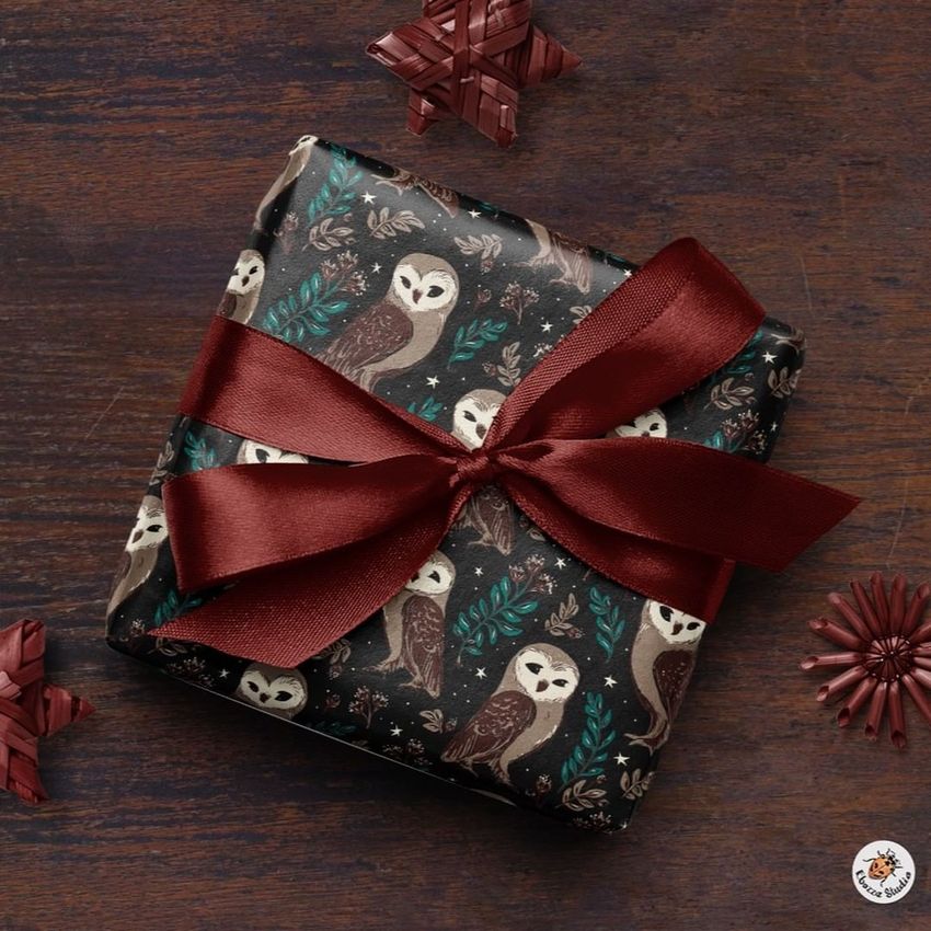 Square gift box mockup wrapped in dark paper with owl and foliage pattern, tied with a burgundy ribbon, on a wooden surface.