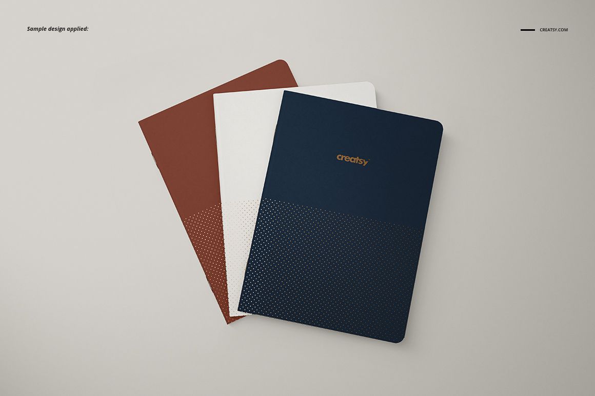 A5 notebook PSD mockup showing three closed notebooks with solid color covers and subtle dotted designs in a top view.