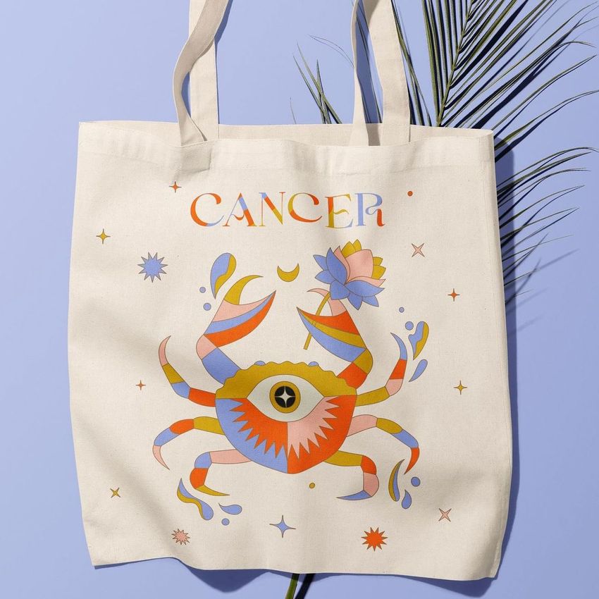 Tote bag mockup featuring a colorful crab illustration and the word 'Cancer' above, displayed on a light purple background with a palm leaf shadow.