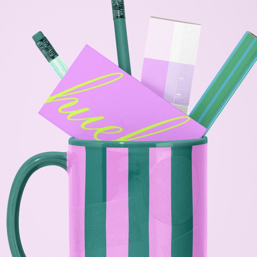 Mug mockup with bold green and pink stripes, filled with office supplies like pencils, a ruler, and purple notecards on a light background.