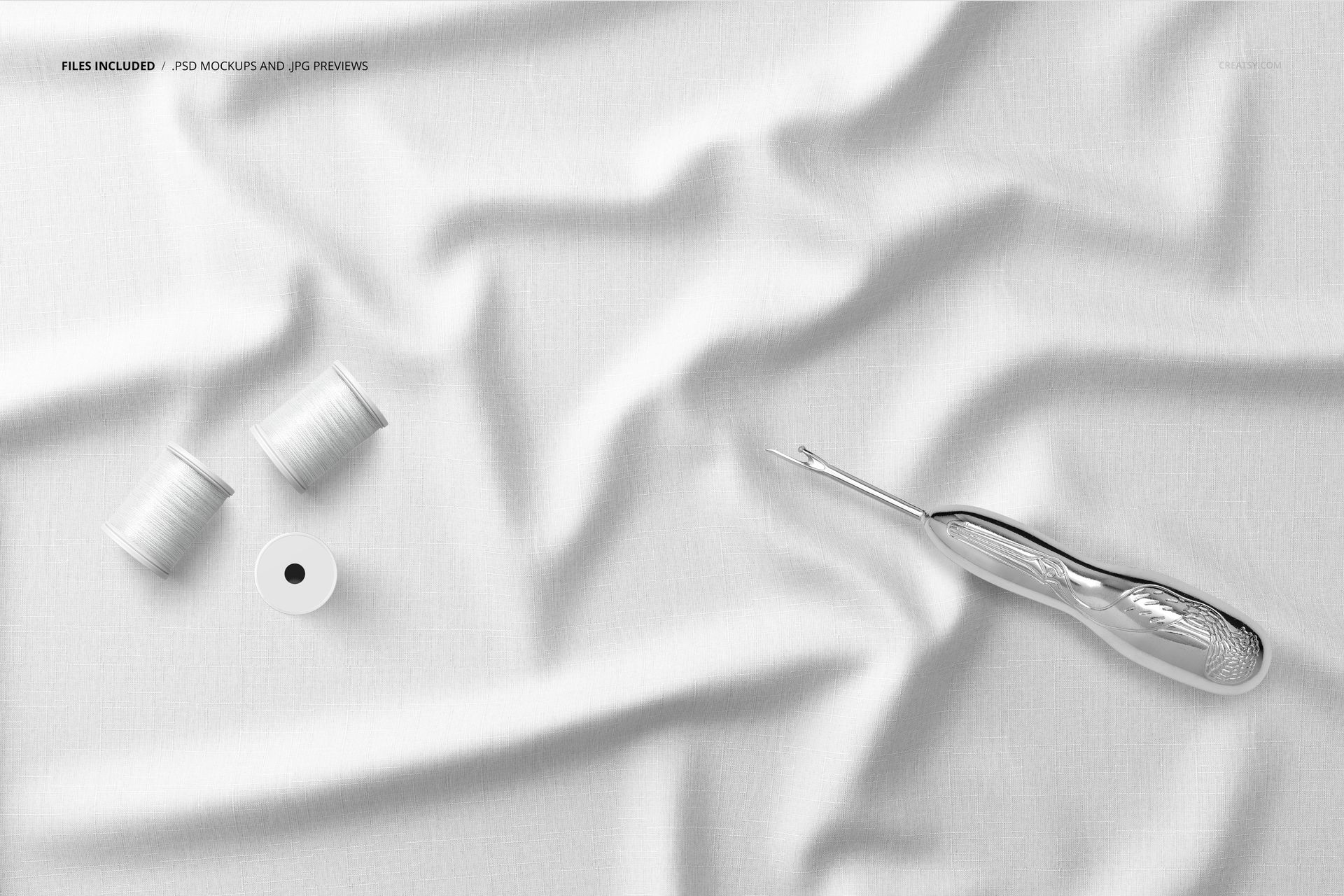 White creased fabric mockup with two spools of thread and a seam ripper, viewed from above on a flat surface.