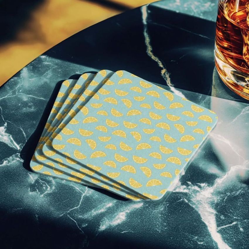Square coaster mockup showing a stack of coasters with yellow lemon slice patterns on a dark marble table in sunlight.