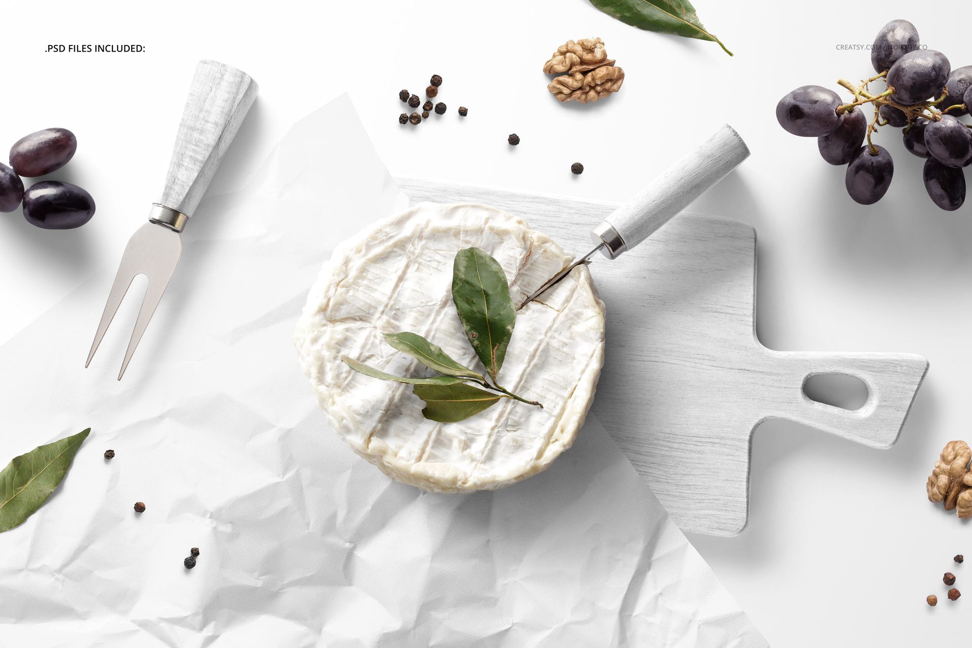 Camembert cheese mockup scene featuring white wrapping paper, wooden board, cheese knife, grapes, walnuts, and green leaves.