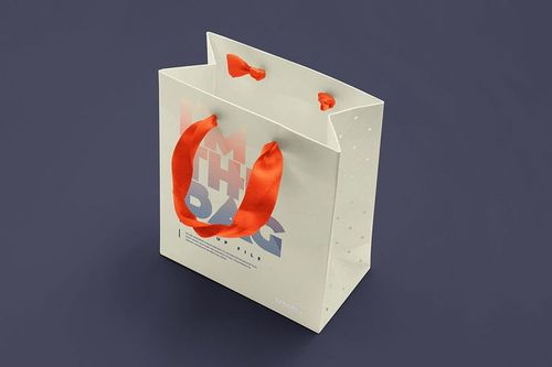 Square gift bag mockup with bold red handles, white body, and modern graphic print, displayed on a neutral dark surface.