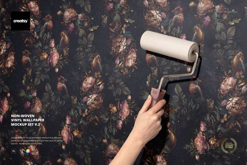 Close-up of a person using a roller to smooth dark floral non-woven vinyl wallpaper in a realistic mockup scene.