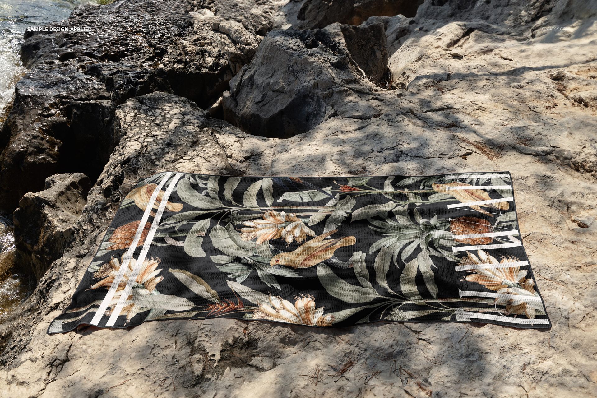 Tropical patterned waffle beach towel mockup displayed on sandy and rocky shore, top-down perspective.