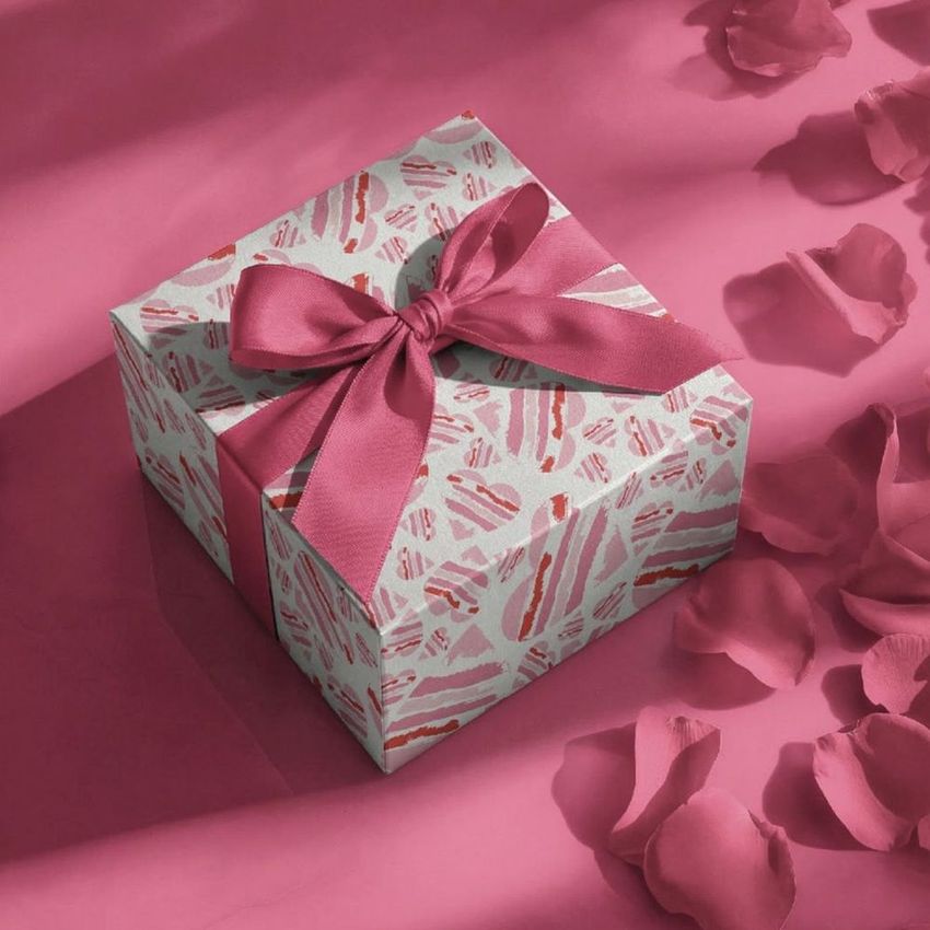 Square gift box mockup with a pink patterned design and a large satin ribbon, surrounded by scattered rose petals on a pink surface.