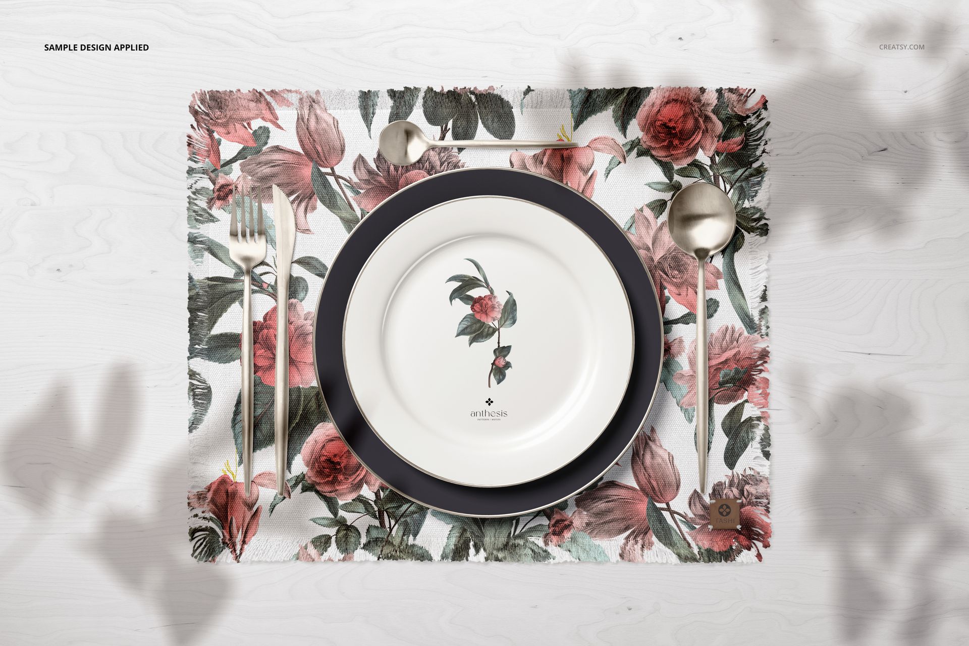 Fringed placemat mockup featuring a floral design, round plates, and silver cutlery arranged on a white wood table.