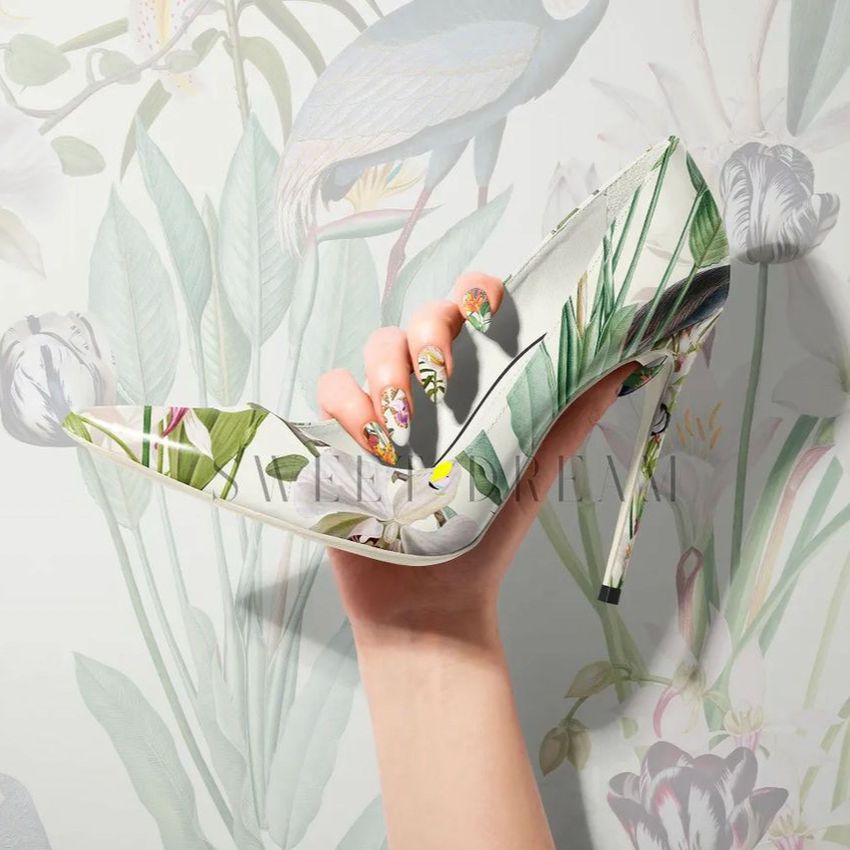 High heels mockup featuring a hand holding a pointed shoe with a botanical pattern, set against a matching floral wallpaper.