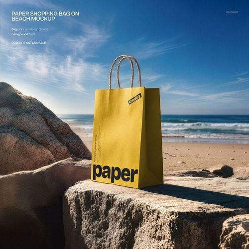 Paper shopping bag mockup in yellow with bold lettering, placed on rocks with beach and ocean scenery in the background.