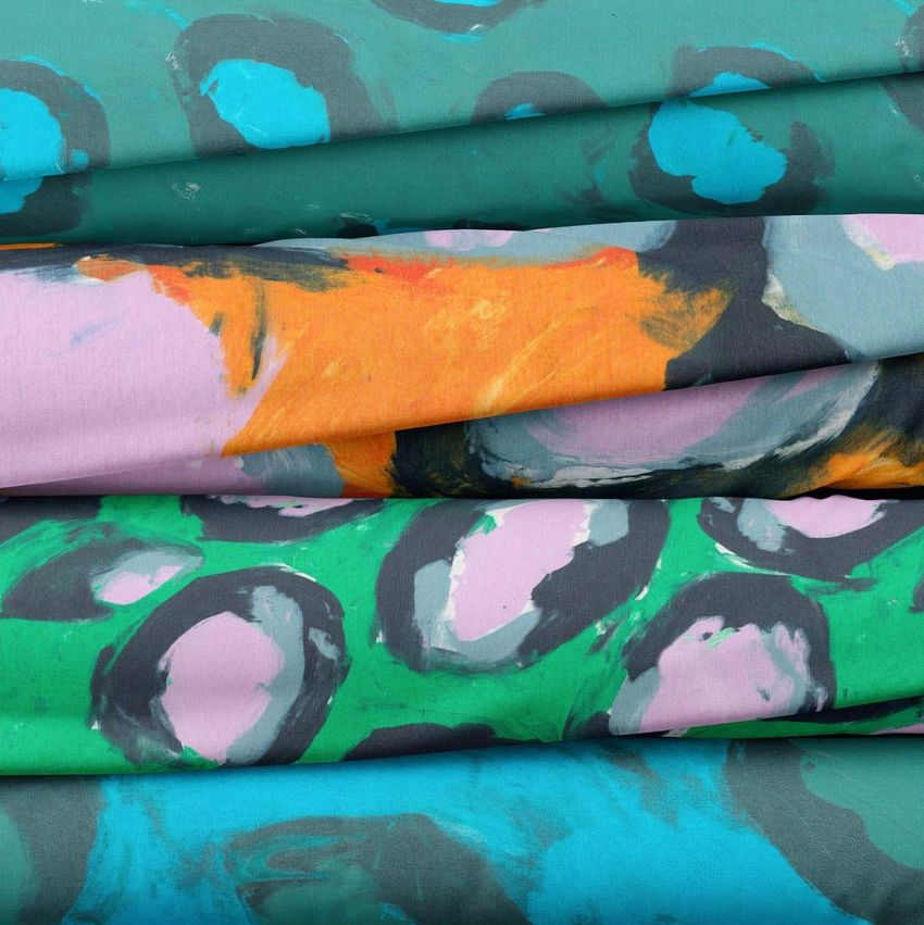 Fabric stack mockup showing folded textiles with abstract, hand-painted patterns in teal, orange, green, and pink hues.