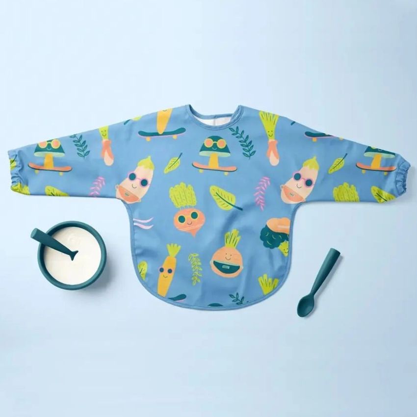 Baby sleeved bib mockup featuring a blue bib with colorful cartoon vegetables and leaves, displayed flat on a light blue surface with a bowl of milk and spoon.