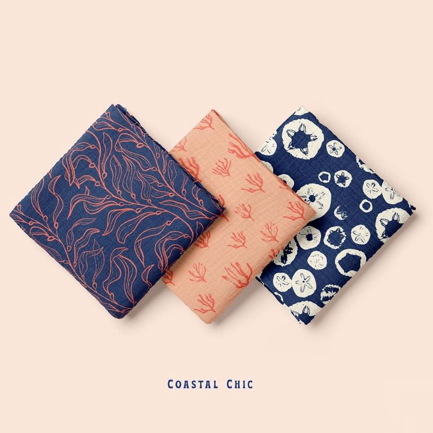 Three folded muslin swaddle blankets with coastal-themed patterns in navy blue, coral, and cream, arranged on a light beige background.