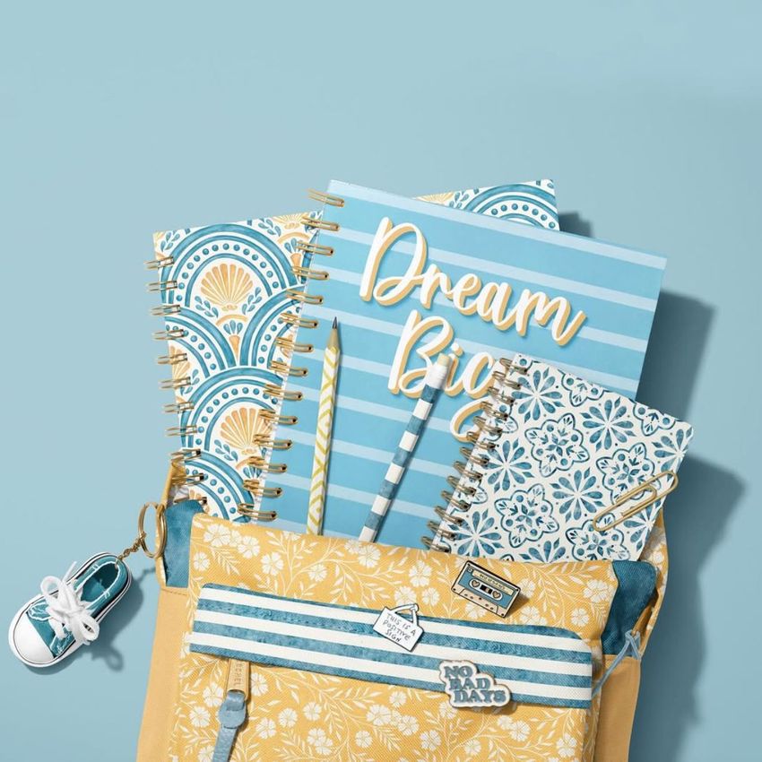 School backpack mockup with spiral notebooks in blue and yellow patterns, a pen, and a small sneaker keychain on a blue background.