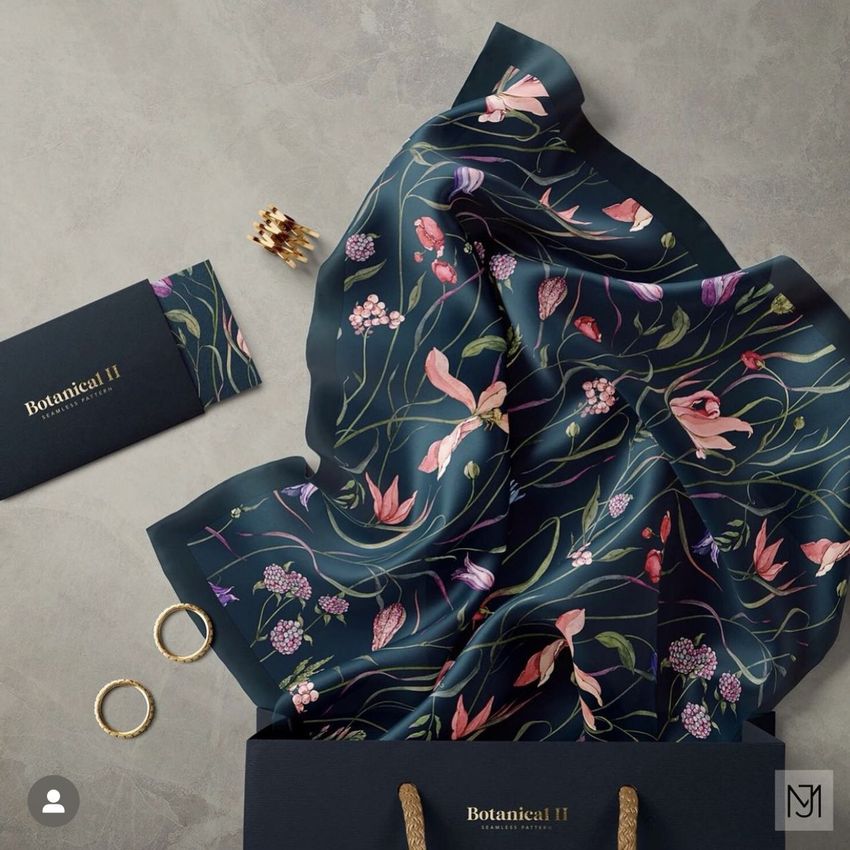 Silk scarf mockup featuring a dark fabric with colorful botanical patterns, displayed partially draped over a shopping bag on a neutral surface.