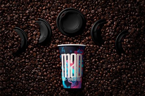 PSD mockup showing a paper cup with a bold graphic design, set in a bed of coffee beans with loose lids nearby.