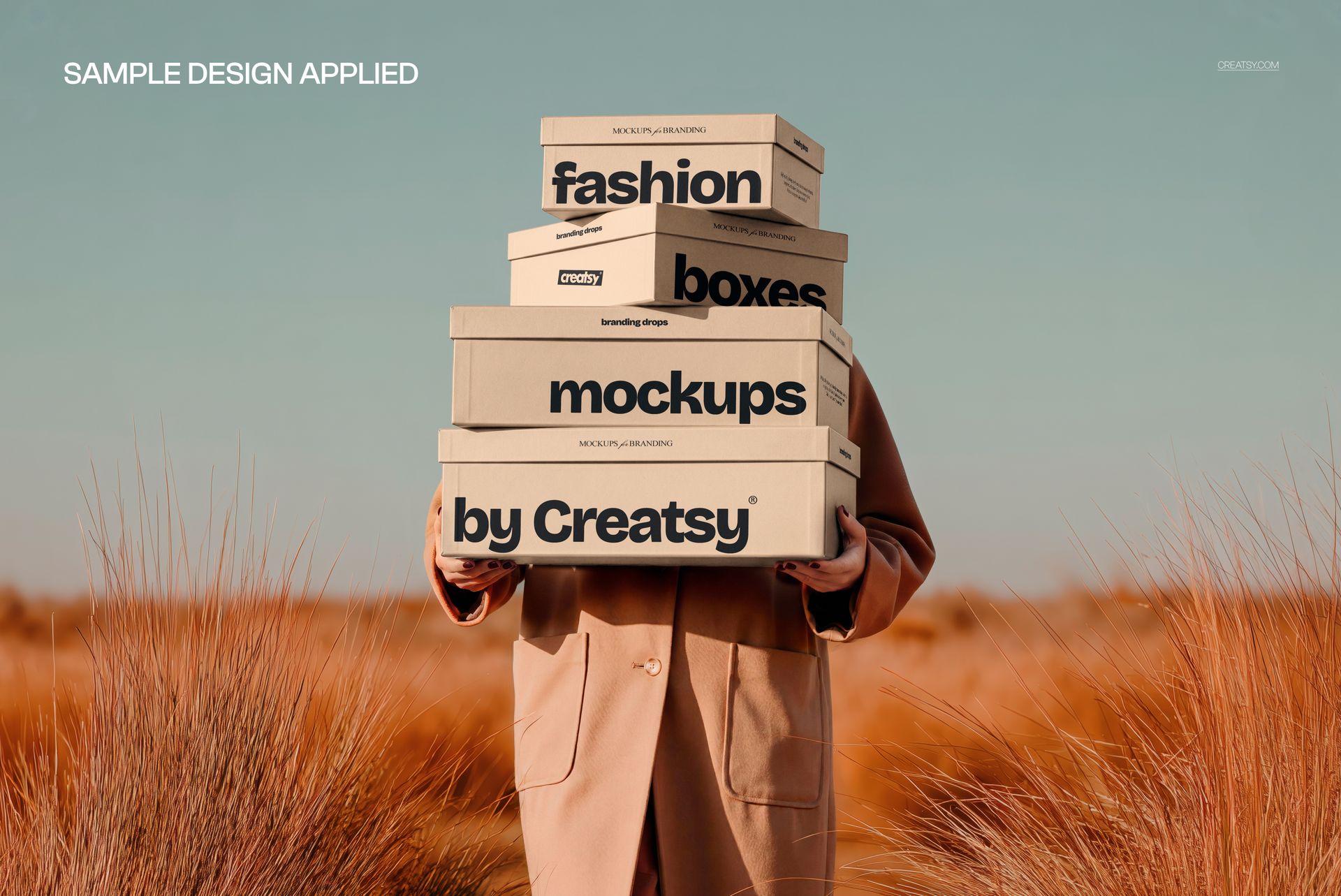 Woman in a tan coat holds four labeled shoe boxes mockup outdoors among tall pampas grass, front perspective.