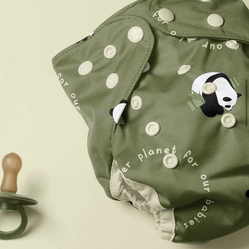 Reusable baby cloth diaper mockup with a green fabric featuring panda and bamboo illustrations, shown next to a pacifier on a light background.
