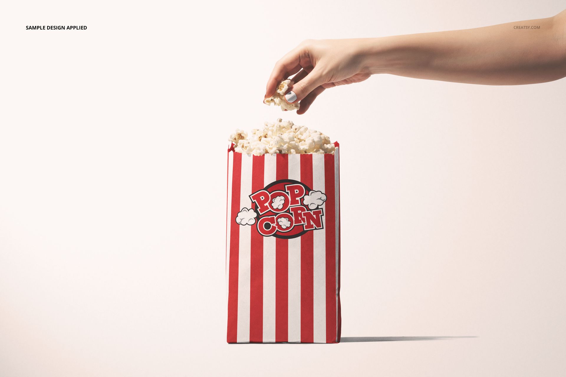 Popcorn paper bag mockup with vertical red and white stripes, overflowing with popcorn, hand grabbing popcorn.