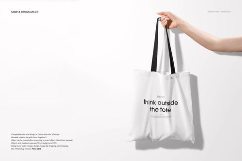 Front-facing mockup of a white tote bag with black straps, displayed in a minimalist setting and held by a hand.