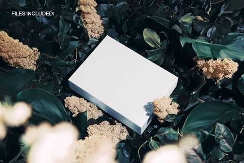 PSD mockup of a blank rectangular box lying amid green foliage and pale flowers, top-down perspective.