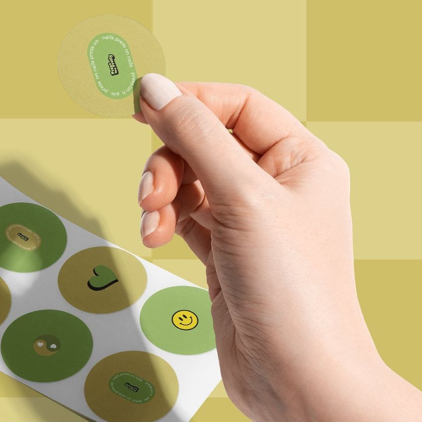 Hand holding a round sticker above a sheet of circle labels, featuring green and yellow colors on a checkered background.