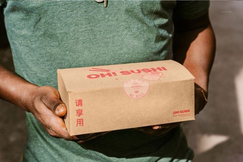 Rectangular takeaway box mockup held in hands outdoors, featuring red sushi-themed graphics on brown cardboard.