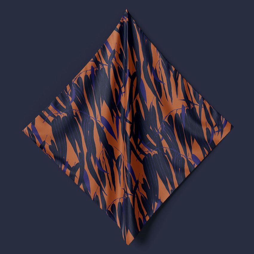 Square silk scarf mockup hanging on a dark wall, featuring an orange and navy abstract floral pattern with soft folds.