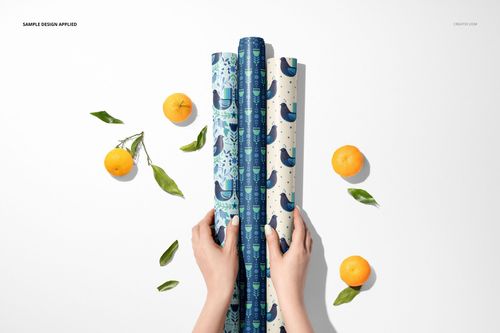 Top view of two hands holding three rolled gift wrapping papers with blue bird patterns, surrounded by oranges.