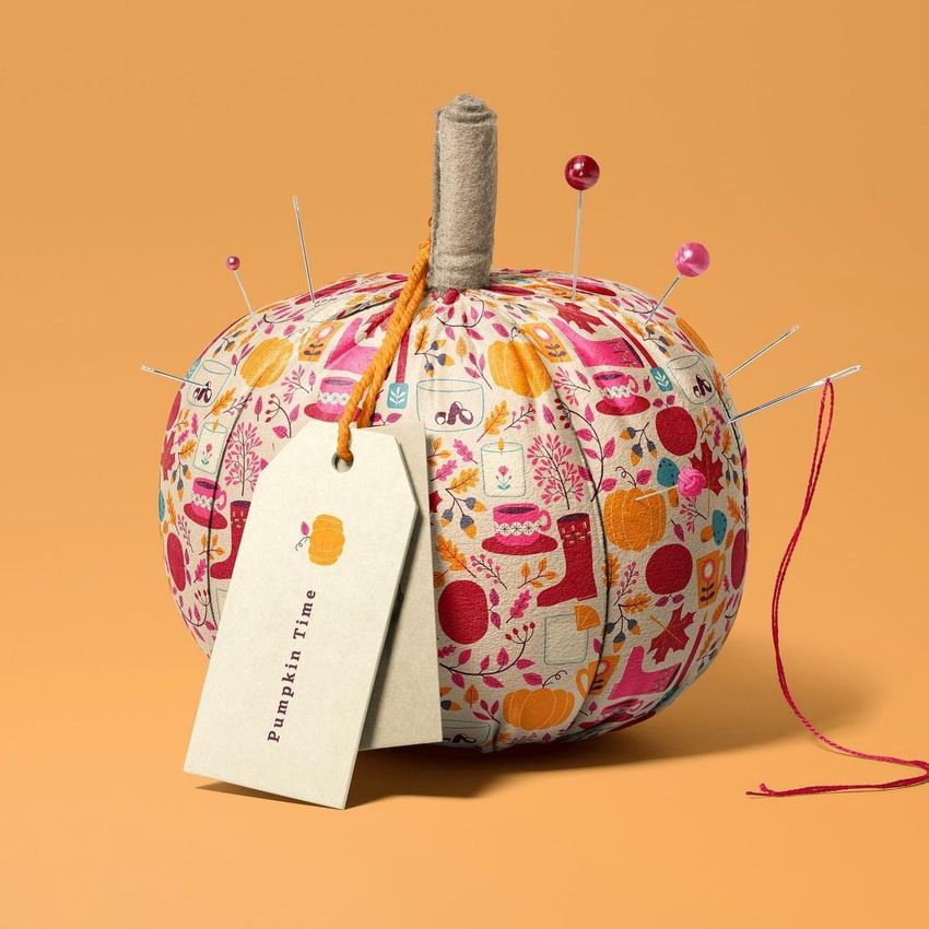 Fabric pumpkin mockup with a colorful autumn-themed pattern, sewing pins, and a tag on an orange background.