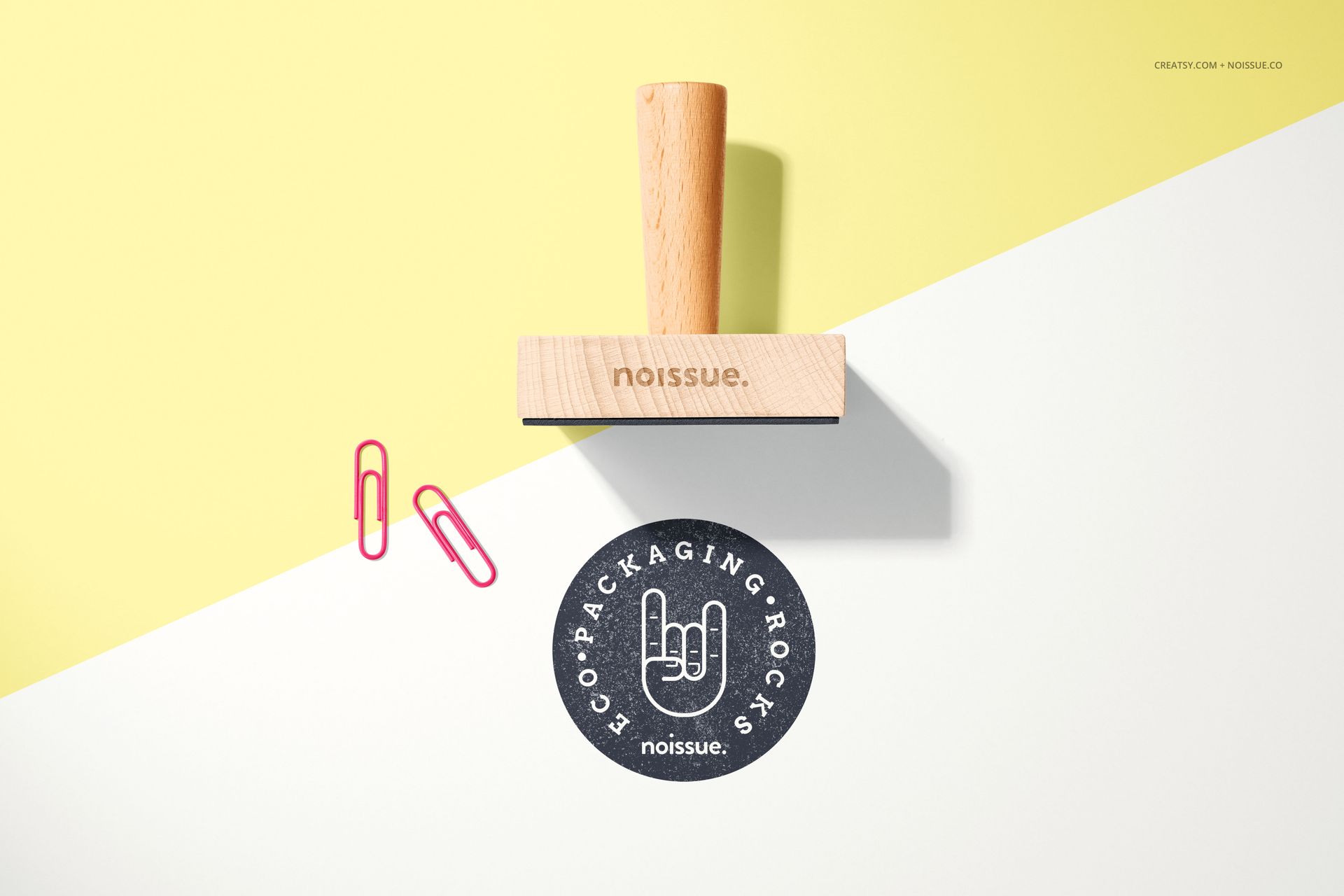 Noissue wooden stamp mockup displayed on a split yellow and white surface with two pink paper clips and a stamped design.