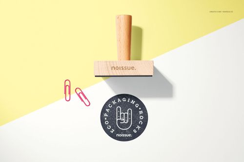 Noissue wooden stamp mockup displayed on a split yellow and white surface with two pink paper clips and a stamped design.