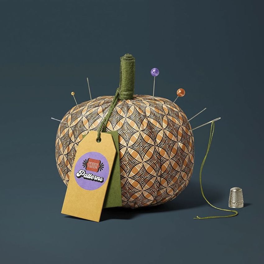 Fabric pumpkin mockup with a geometric brown and orange pattern, used as a pincushion with pins and a tag, on a dark background.