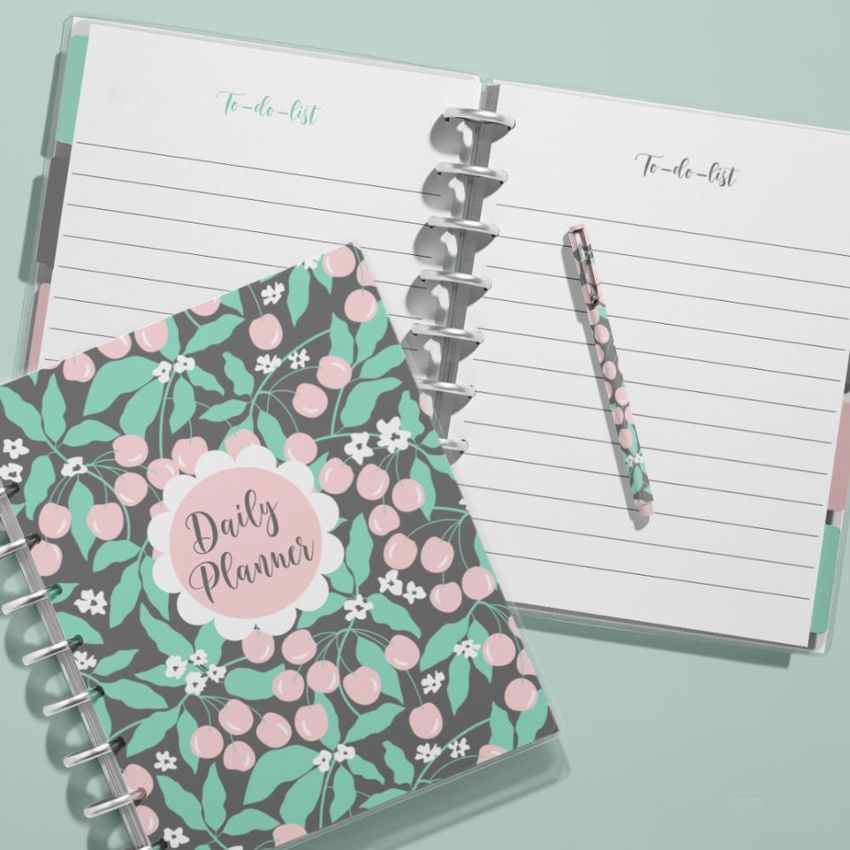Top view of two spiral planners on a light mint background, one open with lined pages and a pen, the other closed with a pink cherry pattern cover.