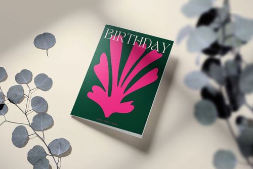 Greeting card mockup featuring a green card with pink abstract artwork, displayed on a neutral surface with eucalyptus leaves.