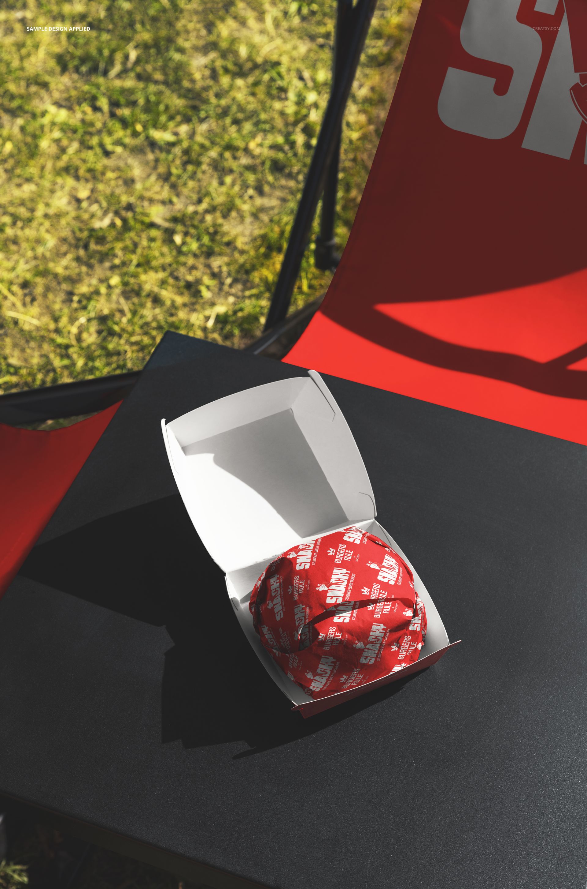 Outdoor setting featuring a red garden folding chair and a black table with a burger mockup in a white box, top view.