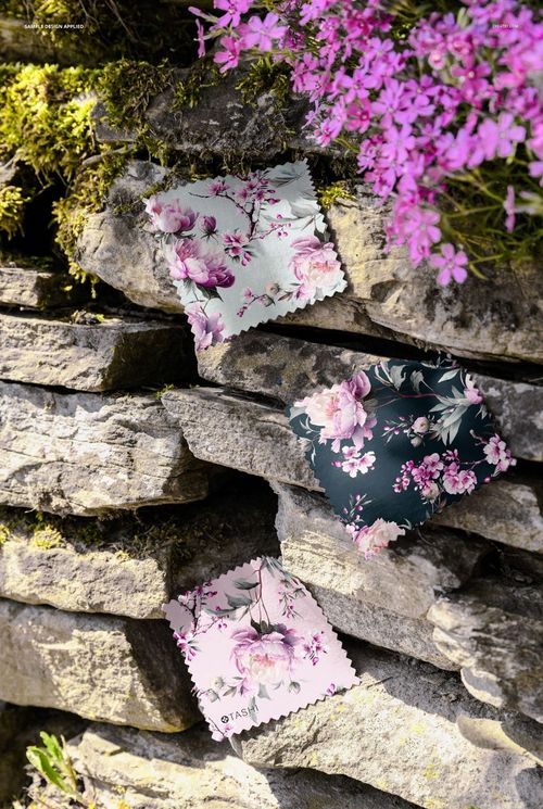 PSD mockup featuring patterned fabric samples placed on moss-covered rocks beneath blooming purple flowers, front view.