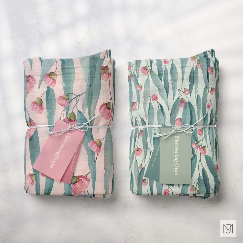 Two muslin swaddle blankets with a flowering gum pattern are folded and tied with string, shown from above on a light surface.